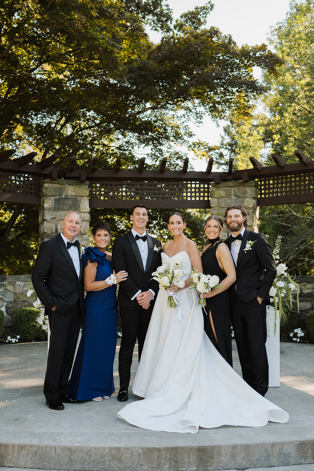 Bride and groom posing for a formal wedding portrait with family under a wooden garden pergola.